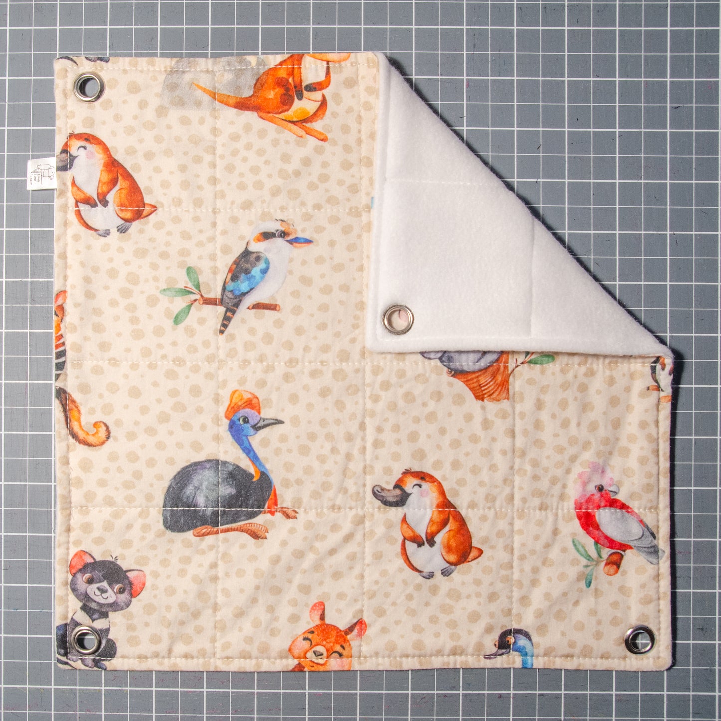 Photo of flat lay flat style hammock with fabric print showing a pale brown background with cartoon characters depicting koalas, platapus, tasmanian devils, emus, kookaburras, numbats and galashs.