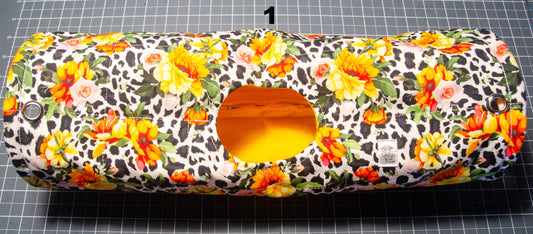 A cylindrical fabric pet bed with a colorful floral and leopard print pattern, featuring a circular opening at the center showing a yellow interior. The item is placed on a black and white grid cutting mat.